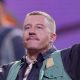 Close up of Macklemore, who has shaved blonde hair, and is wearing a blue striped shirt.