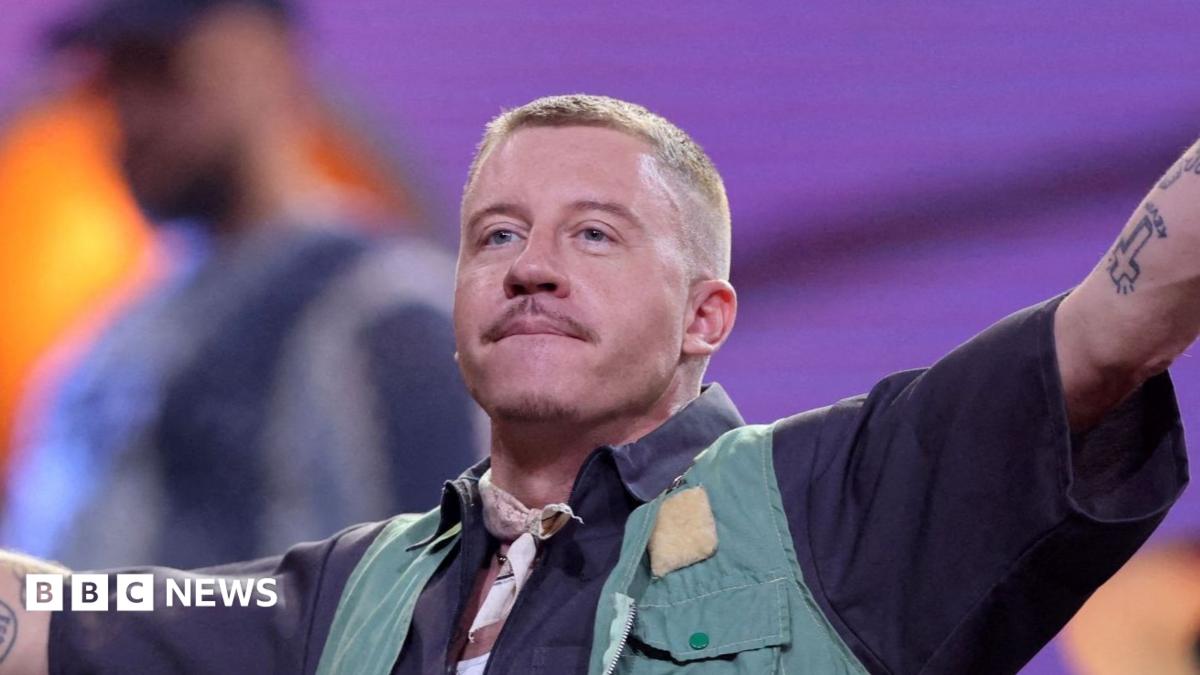 Close up of Macklemore, who has shaved blonde hair, and is wearing a blue striped shirt.