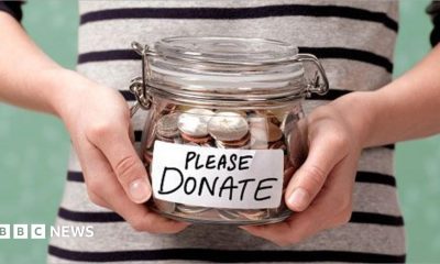 A shot of someone's torso, in a black and white striped top, holding a glass jar with coins in it. On the front is a label that reads: Please donate.