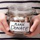 A shot of someone's torso, in a black and white striped top, holding a glass jar with coins in it. On the front is a label that reads: Please donate.