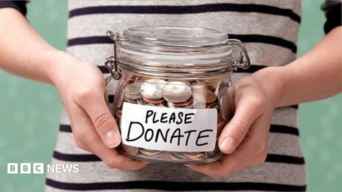 A shot of someone's torso, in a black and white striped top, holding a glass jar with coins in it. On the front is a label that reads: Please donate.