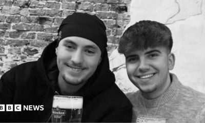 A black and white photo of Eddie and Jayden, smiling side by side. They are both holding pints or beer. Jayden has a hat on and has short stubble and is wearing a black hoodie. Eddie has short dark hair and is wearing a jumper.