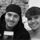 A black and white photo of Eddie and Jayden, smiling side by side. They are both holding pints or beer. Jayden has a hat on and has short stubble and is wearing a black hoodie. Eddie has short dark hair and is wearing a jumper.