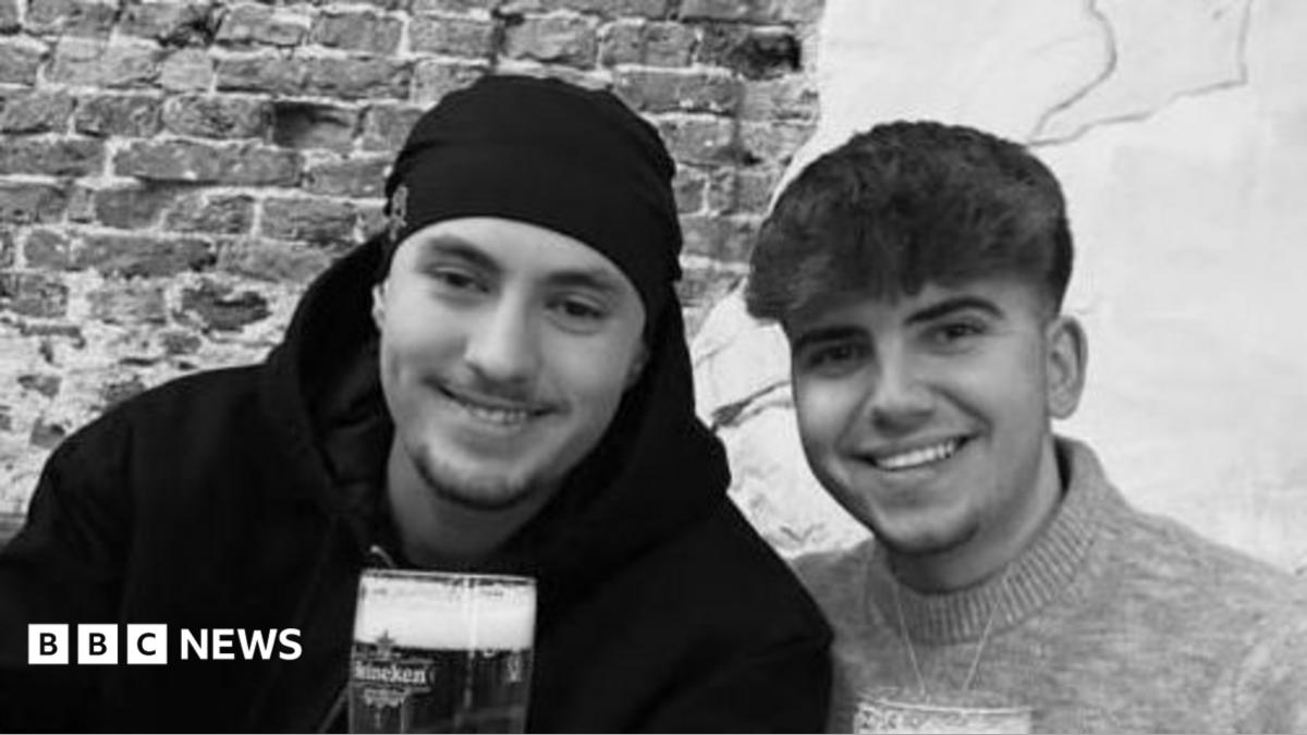A black and white photo of Eddie and Jayden, smiling side by side. They are both holding pints or beer. Jayden has a hat on and has short stubble and is wearing a black hoodie. Eddie has short dark hair and is wearing a jumper.