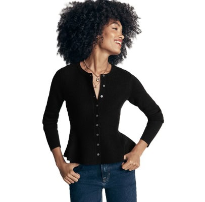 A woman wearing a black peplum cardigan, blue jeans, and gold necklaces