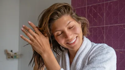 Portrait of young beautiful woman in a bathroom performing cosmetic procedures after bathing. Female wearing bathrobe, applying haircare product and smiling. Close up, copy space background.