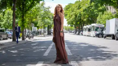 Chloé Lecareux wears a halter-neck dress in a rich brown satin fabric, featuring a flowing silhouette. The dress is complemented by a gold metallic collar detail. Hair is styled in loose waves. Accessories include a chunky bracelet with large, dark stones set in gold frames. A textured sequined brown handbag is carried, outside Stephane Rolland, during Paris Fashion Week - Haute Couture Fall/Winter 2025/2026, on July 08, 2025 in Paris, France
