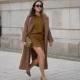 Laura Noltemeyer is seen wearing a a long camel-colored brushed wool coat with a wrap silhouette and integrated belt by Max Mara; a mustard-colored knit cardigan with a brushed cable-knit pattern and front button fastening by Sézane; a mustard yellow brownish mini skirt in a soft wool fabric by Sézane; black patent leather slingback pumps with a sharply pointed toe and silver hardware accents by Yves Saint Laurent; a small quilted tweed flap bag in a khaki Color with woven fabric material with a gold chain strap and CC clasp by Chanel; gold statement earrings with a ruffled surface and a green stone element by Mango; black cat eye sunglasses with an acetate frame by Yves Saint Laurent; her brown hair is styled in loose waves with a center part on November 13, 2025 in Hamburg, Germany.