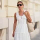 Diane Batoukina wears brown hair with a center part pulled back into a low bun, black rectangular aviator sunglasses with dark lenses by Kaleos, an ivory white sleeveless midi dress with a scoop neck, lace trim, broderie anglaise, floral embroidery and tiered gathered panels by Rouje, a cream white woven tote bag with brown leather shoulder straps by Liffner, during a street style fashion photo session, on May 26, 2026 in Paris, France.