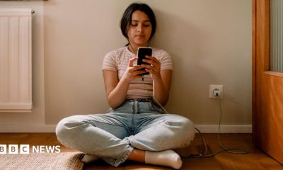 A young girl is seen sat crossed legged on the floor scrolling on a smartphone while it is charging