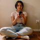 A young girl is seen sat crossed legged on the floor scrolling on a smartphone while it is charging