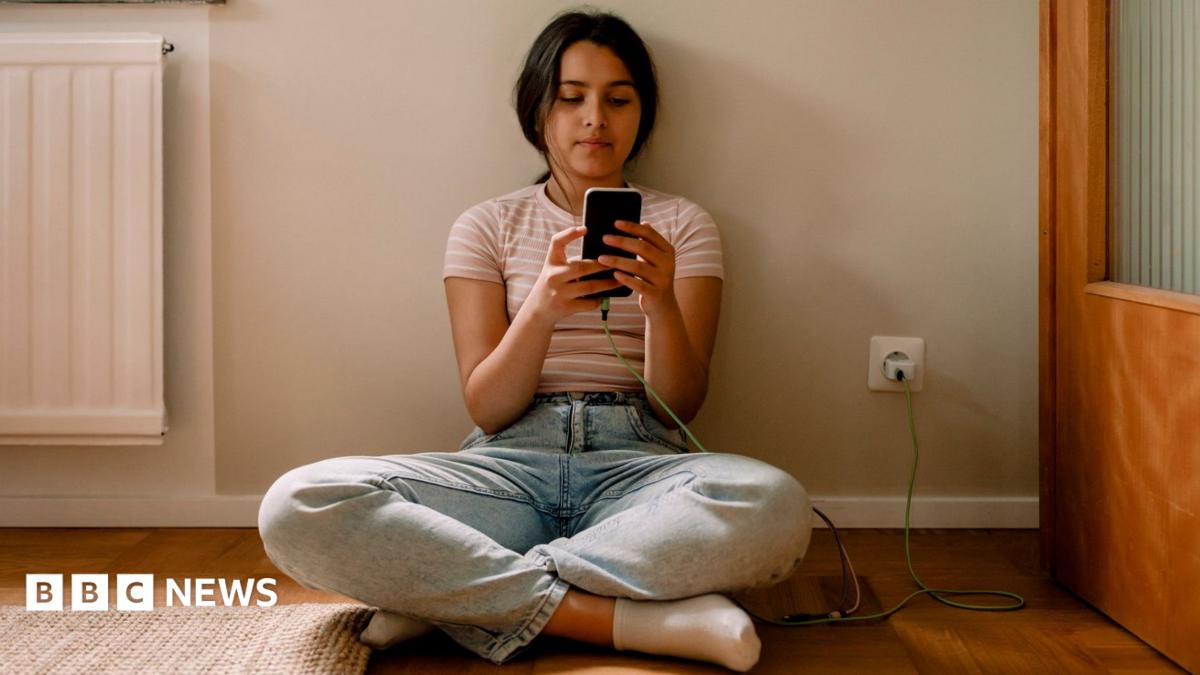 A young girl is seen sat crossed legged on the floor scrolling on a smartphone while it is charging
