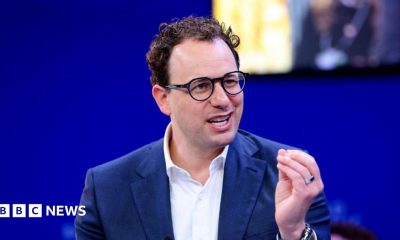 Dario Amodei hld the closed fingers of his left hand together as he mades a point. He is wearing a white shirt and blue jacket, black-rimmed glasses and has slightly curly hair. He is sat against a blue background, with a yellow-lit square just behind and to the left of his head.