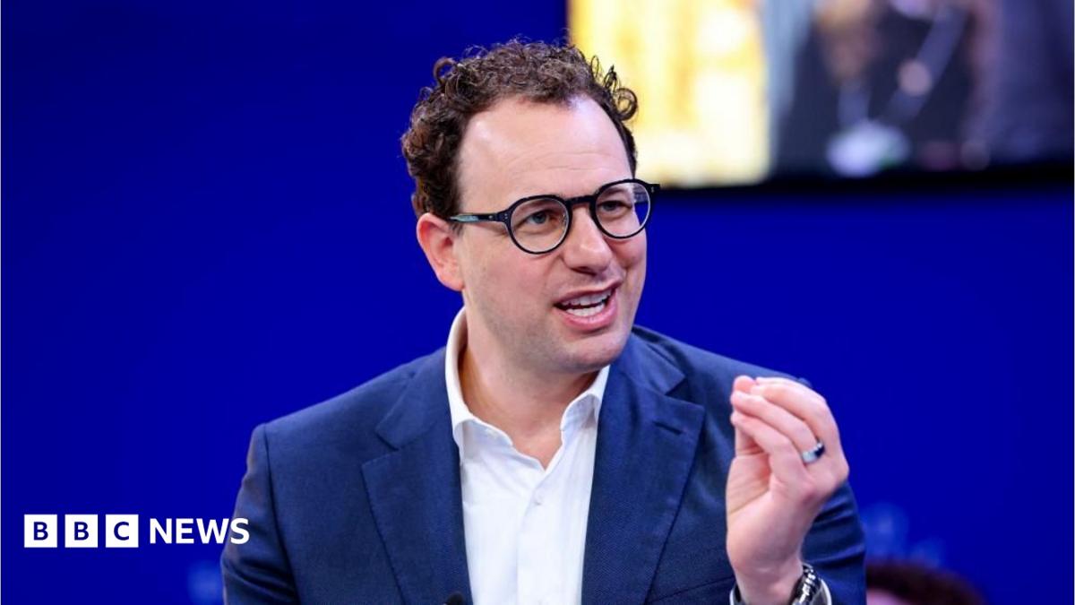 Dario Amodei hld the closed fingers of his left hand together as he mades a point. He is wearing a white shirt and blue jacket, black-rimmed glasses and has slightly curly hair. He is sat against a blue background, with a yellow-lit square just behind and to the left of his head.