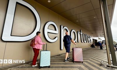 A man and a woman roll pink and blue suitcases past a large "Departures" sign at Belfast airport.