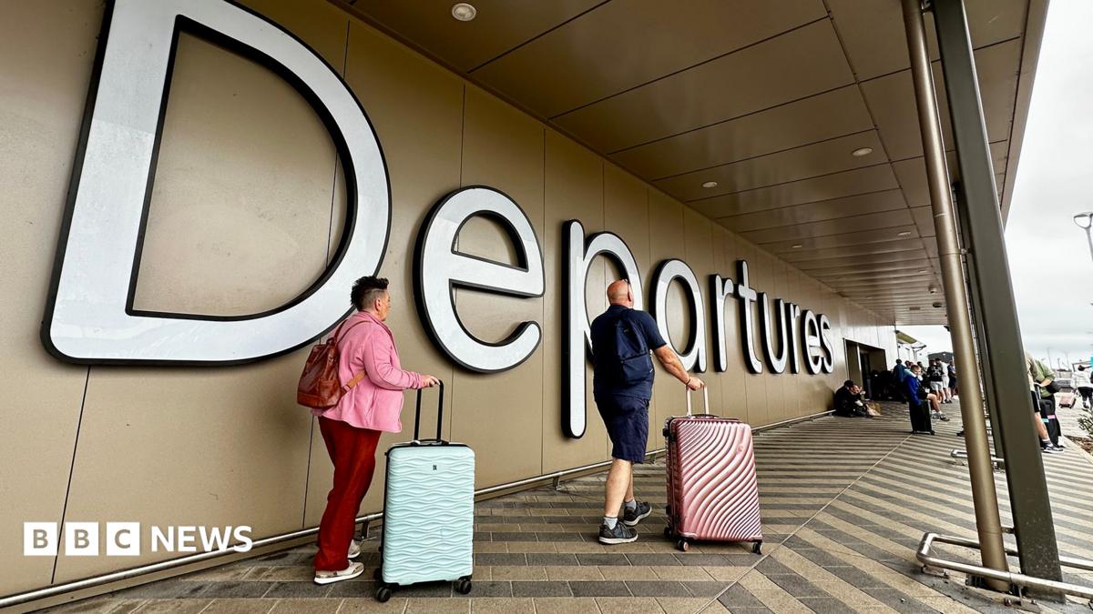 A man and a woman roll pink and blue suitcases past a large "Departures" sign at Belfast airport.