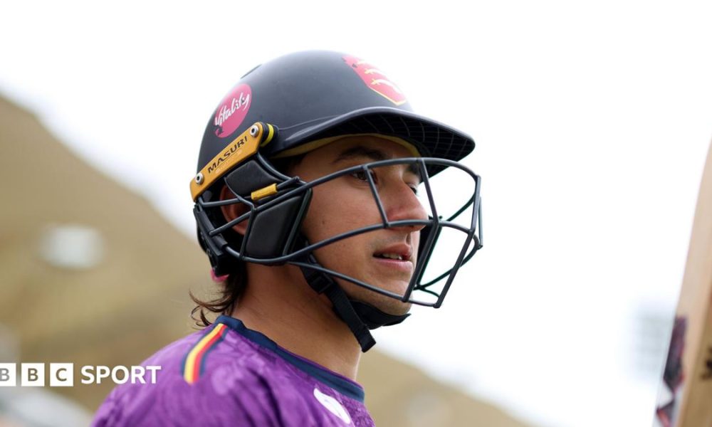 Robin Das wearing a black cricket helmet, carrying a cricket bat and wearing a purple top