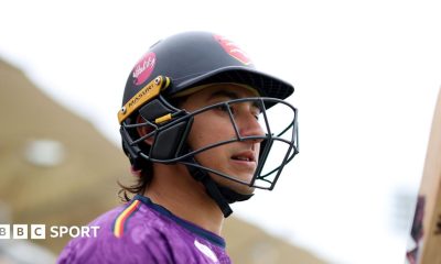 Robin Das wearing a black cricket helmet, carrying a cricket bat and wearing a purple top