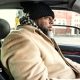 A bearded man in a beige coat and a black and white chequered scarf drives his car through a US urban area. His car has a black leather interior.