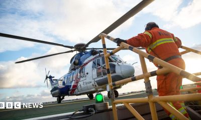 A helicopter lands on an oil rig and a man in orange overalls is climbing up steps and heading towards it