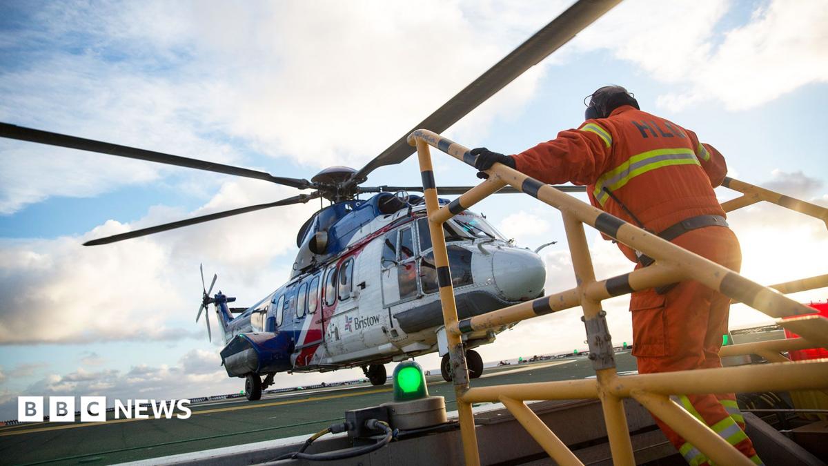 A helicopter lands on an oil rig and a man in orange overalls is climbing up steps and heading towards it