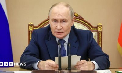 Putin sits in front of a desk and microphone holding some notes