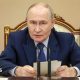 Putin sits in front of a desk and microphone holding some notes