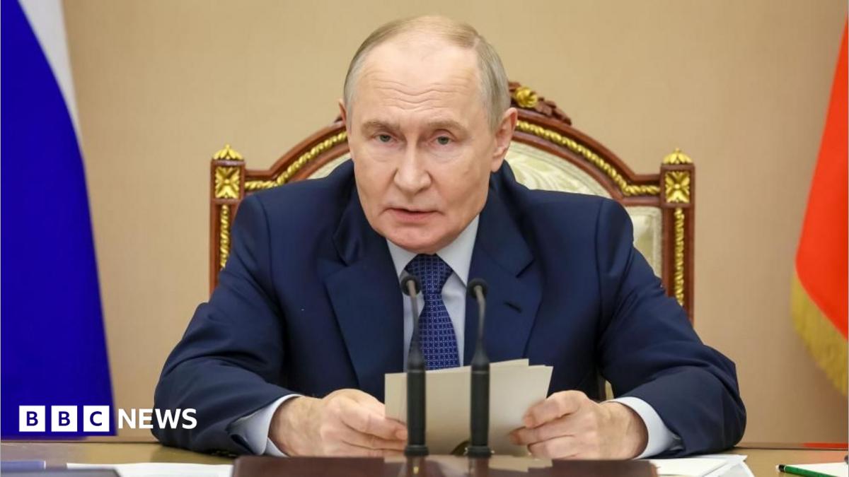 Putin sits in front of a desk and microphone holding some notes