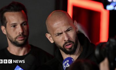 Tristan and Andrew Tate delivering a press statement. The brothers are dressed in black and Andrew has a disgruntled facial expression as he speaks into a microphone.