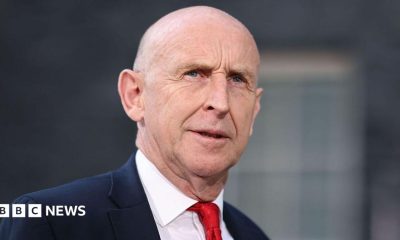 John Healey stands looking into the middle distance wearing a red tie, white shirt and navy blue suit. There is a blurred window behind him.