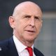 John Healey stands looking into the middle distance wearing a red tie, white shirt and navy blue suit. There is a blurred window behind him.