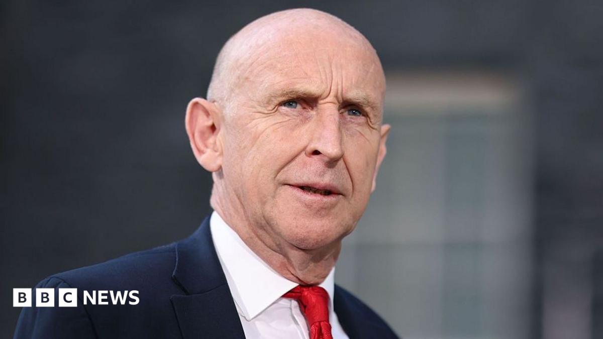 John Healey stands looking into the middle distance wearing a red tie, white shirt and navy blue suit. There is a blurred window behind him.
