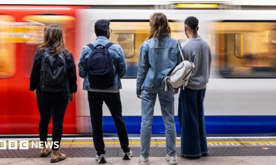 Four young people stand on a London underground platform facing the tracks as a train arrives.