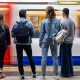 Four young people stand on a London underground platform facing the tracks as a train arrives.