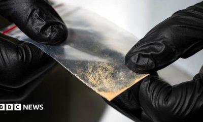 Close-up of gloved hands holding a small transparent plastic packet containing a light-coloured powdery substance. The packet is being examined against an out-of-focus laboratory background.