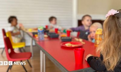 The image shows a group of unidentifiable toddlers sitting around a table eating and drinking. The table is red and the children's faces are blurry.