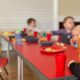 The image shows a group of unidentifiable toddlers sitting around a table eating and drinking. The table is red and the children's faces are blurry.