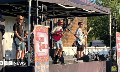 Four men on a small stage in the sun. Three of the men are playing guitars and one is sat at a drum kit. The stage is sheltered by a pop-up gazebo.