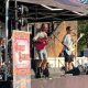 Four men on a small stage in the sun. Three of the men are playing guitars and one is sat at a drum kit. The stage is sheltered by a pop-up gazebo.