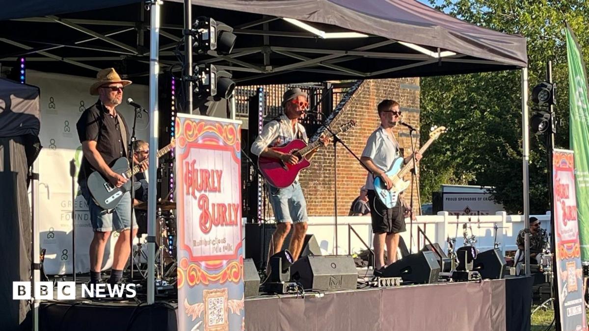 Four men on a small stage in the sun. Three of the men are playing guitars and one is sat at a drum kit. The stage is sheltered by a pop-up gazebo.