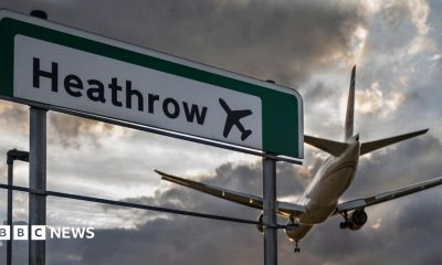 A commercial aeroplane flying low on final approach to land at Heathrow Airport, passing directly over an overhead highway road sign.