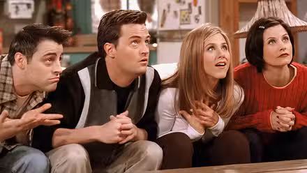 The cast of Friends on a couch looking up at a TV.