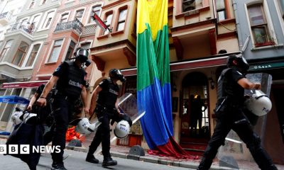 Riot police, with a rainbow flag in the background, chase LGBT activists as they try to gather for a pride parade, which was banned by the governorship in Istanbul. Officers are wearing black uniforms, wearing gas masks, and are carrying helmets and riot shields as they move through what appears to be a residential street.