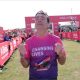 Andy Burnham crossing the finishing line in South Shields. He is clenching his fists and looking upwards with an expression of strain, and wearing a pink and purple T-shirt with changing lives on it. Behind him stretch out the hoardings with crowds of spectators, and another runner leans on it to the left, with two more standing on the right.