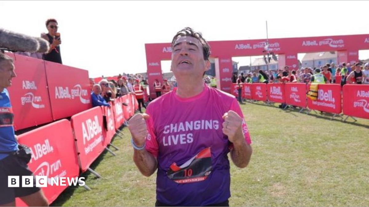 Andy Burnham crossing the finishing line in South Shields. He is clenching his fists and looking upwards with an expression of strain, and wearing a pink and purple T-shirt with changing lives on it. Behind him stretch out the hoardings with crowds of spectators, and another runner leans on it to the left, with two more standing on the right.