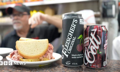 A sandwich and two types of cherry cola on a countertop in a deli.