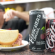 A sandwich and two types of cherry cola on a countertop in a deli.