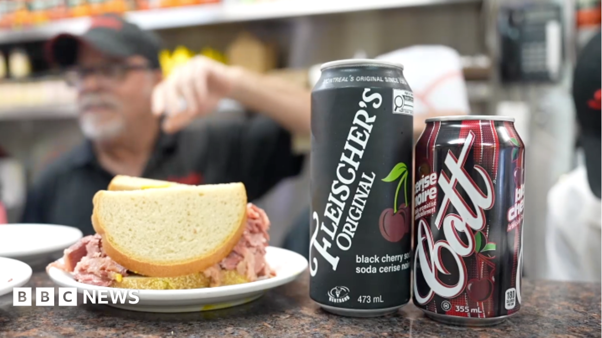 A sandwich and two types of cherry cola on a countertop in a deli.