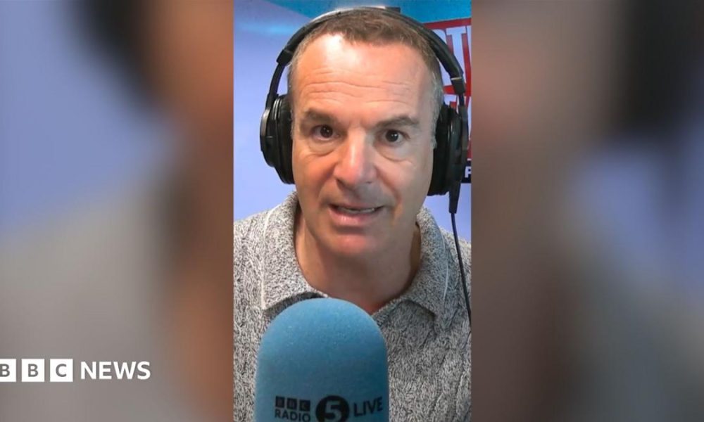 Martin Lewis looks into the camera. He is wearing headphones and is sat in front of a microphone with a blue mic muff. Only his face and shoulders are visible.