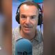 Martin Lewis looks into the camera. He is wearing headphones and is sat in front of a microphone with a blue mic muff. Only his face and shoulders are visible.